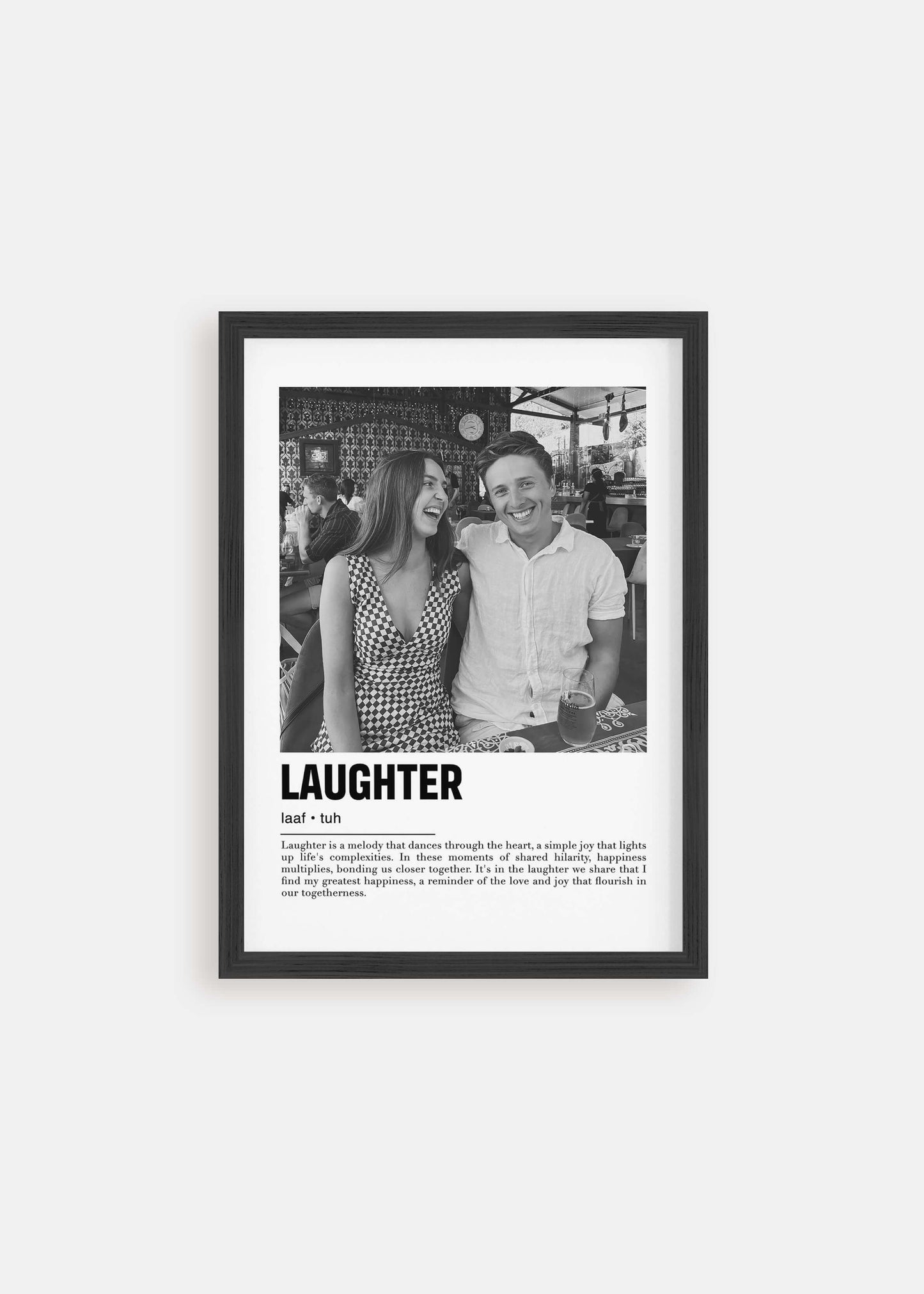 Personalized black and white photo in a black frame of a couple laughing. The custom artwork has a caption that reads Laughter. The black frame is on a white background.