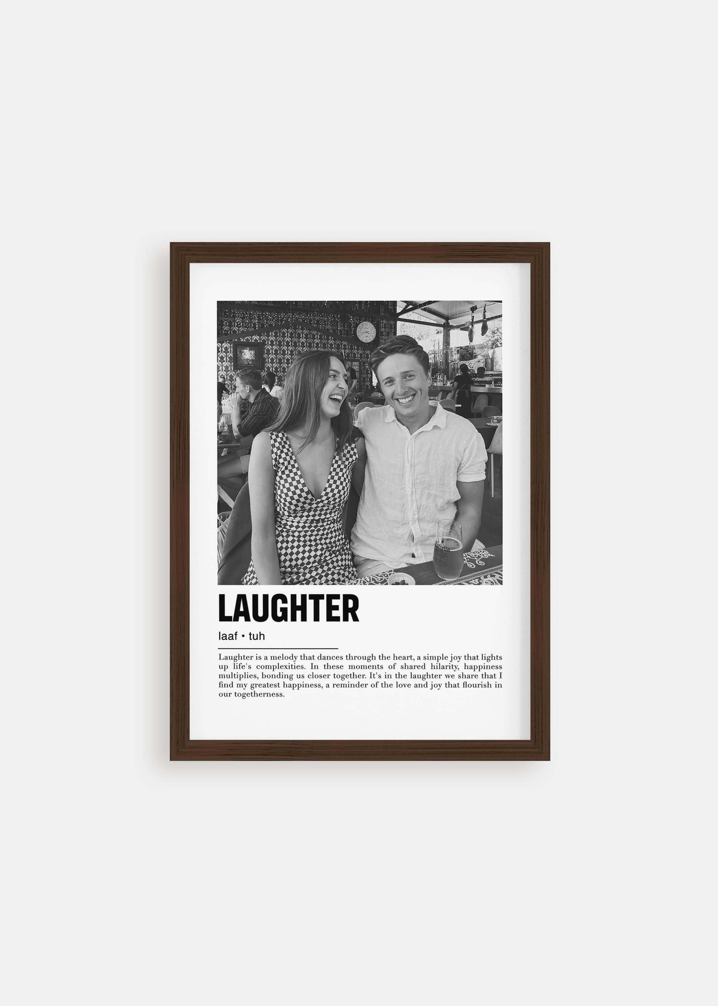 Personalized black and white photo in a walnut frame of a couple laughing. The custom artwork has a caption that reads Laughter. The walnut frame is on a white background.