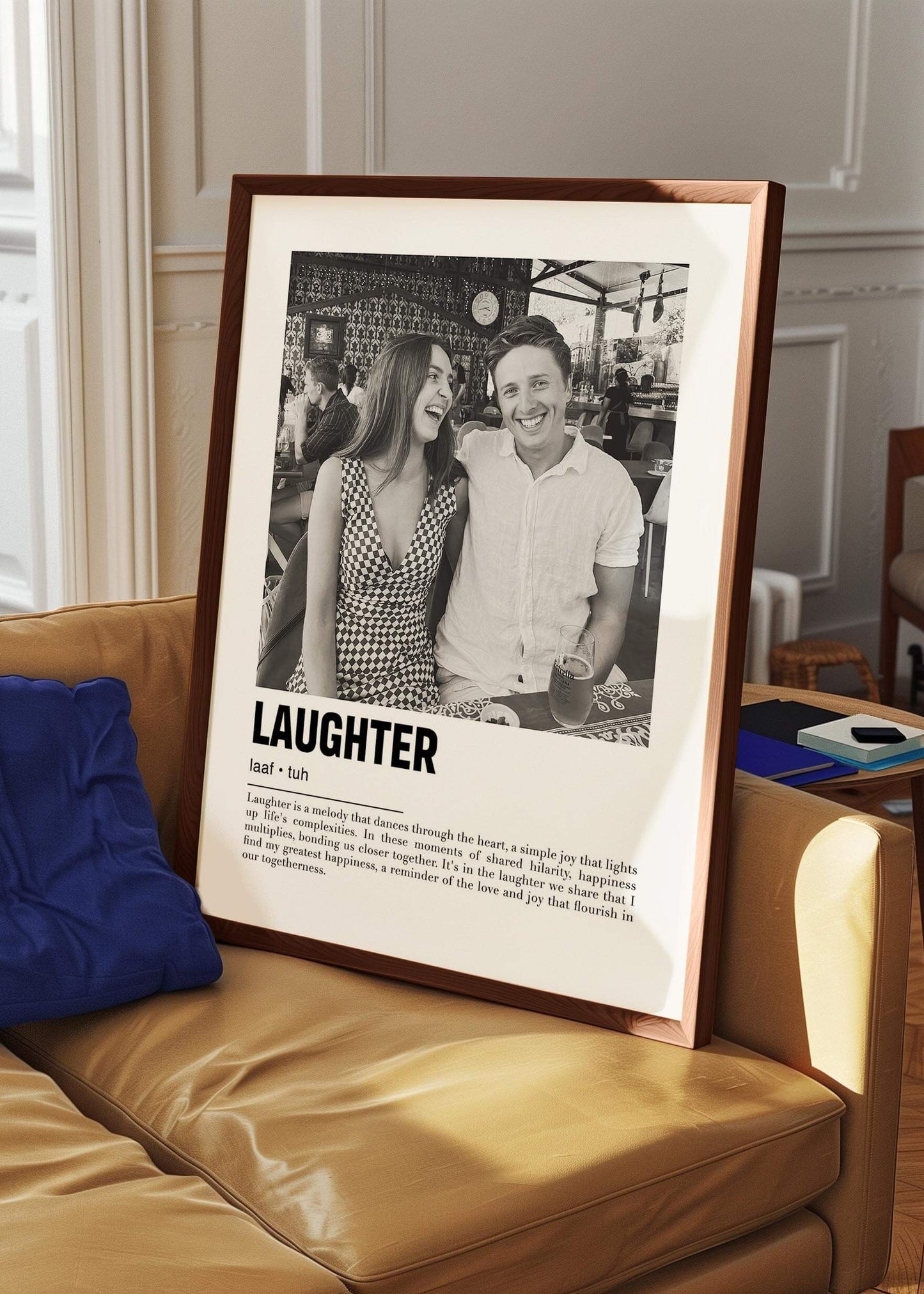 Personalized black and white photo in a walnut frame of a couple laughing. The custom artwork has a caption that reads Laughter. The walnut frame is on a sofa.
