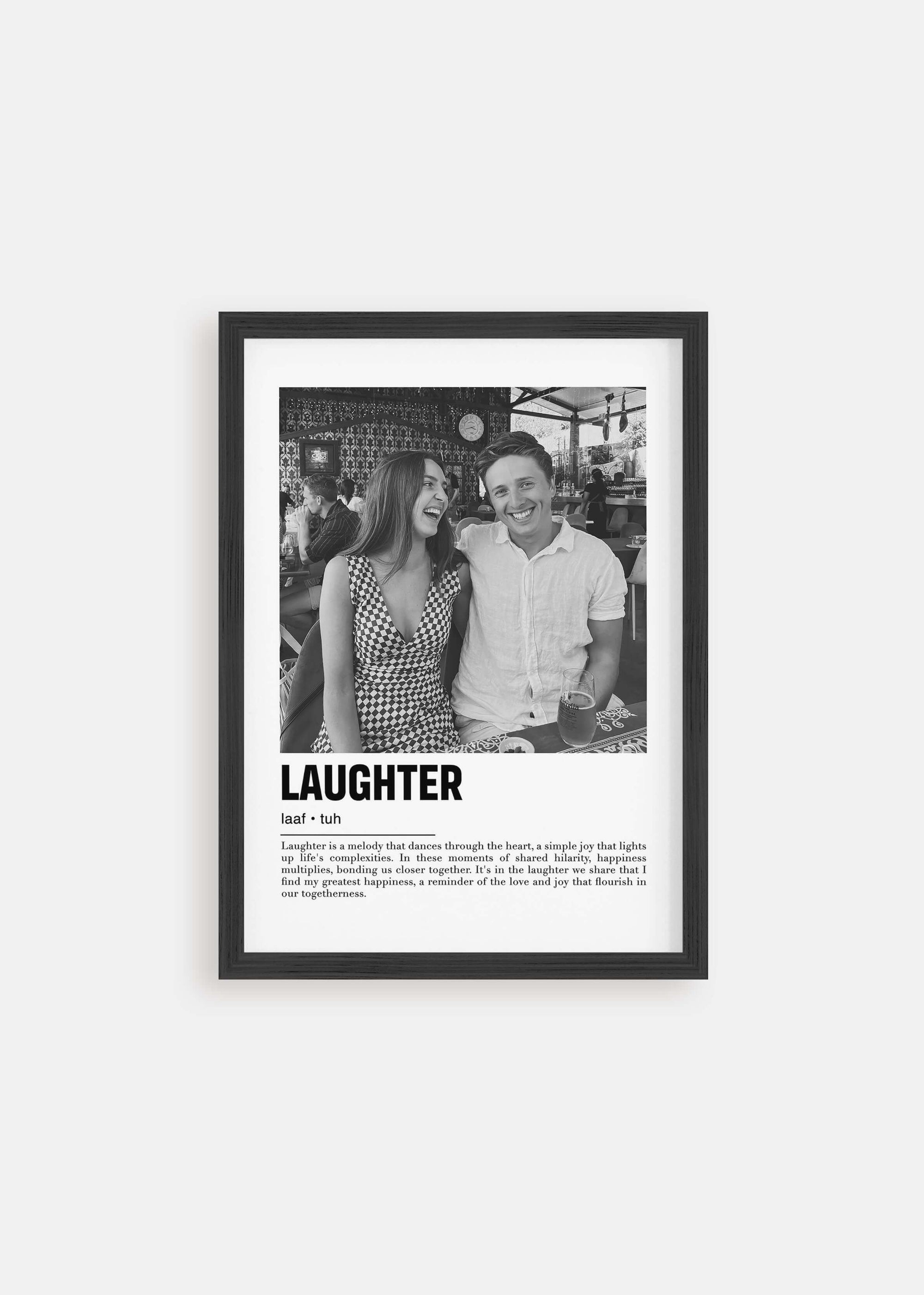 Personalized black and white photo in a black frame of a couple laughing. The custom artwork has a caption that reads Laughter. The black frame is on a white background. 