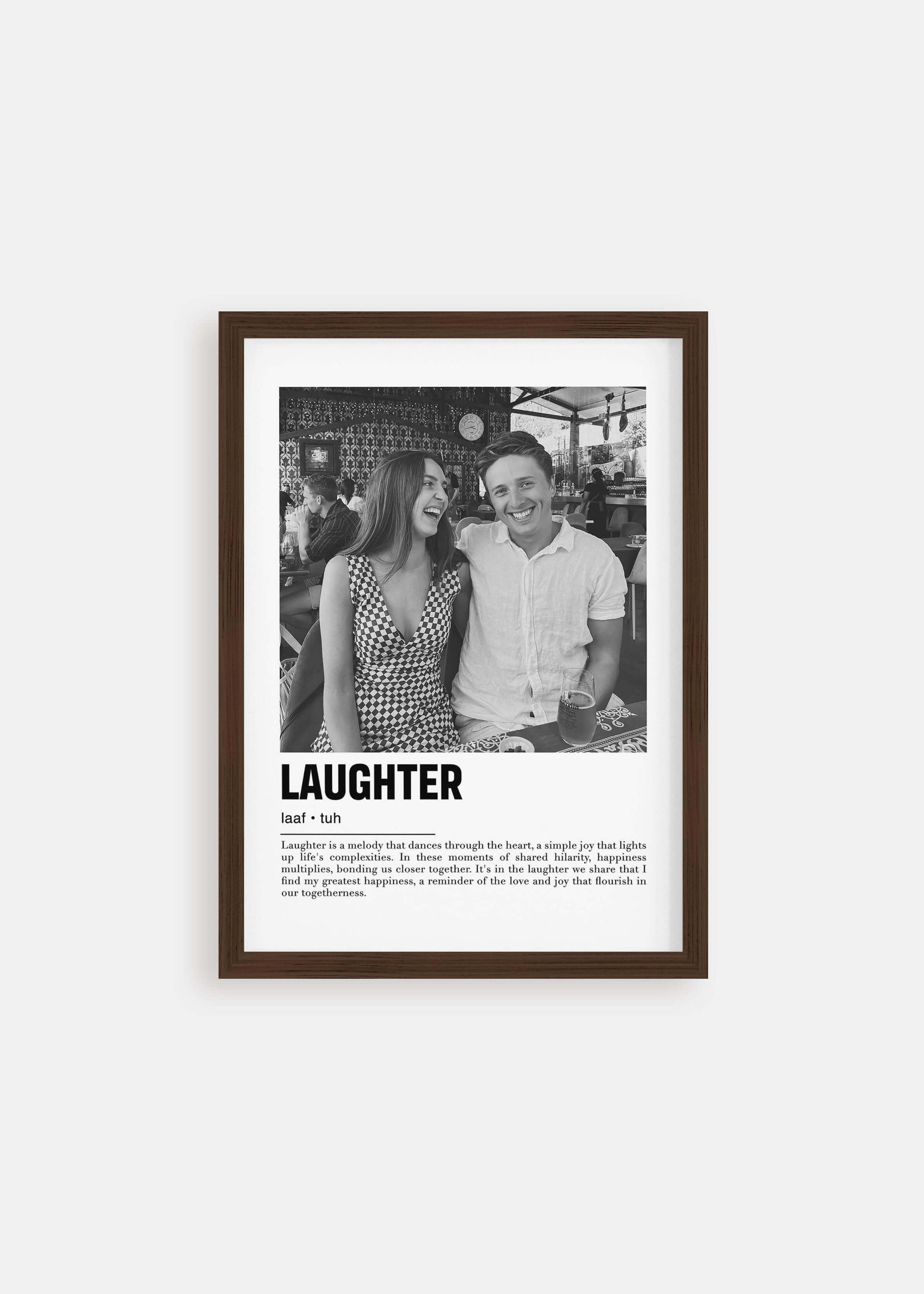 Personalized black and white photo in a walnut frame of a couple laughing. The custom artwork has a caption that reads Laughter. The walnut frame is on a white background. 