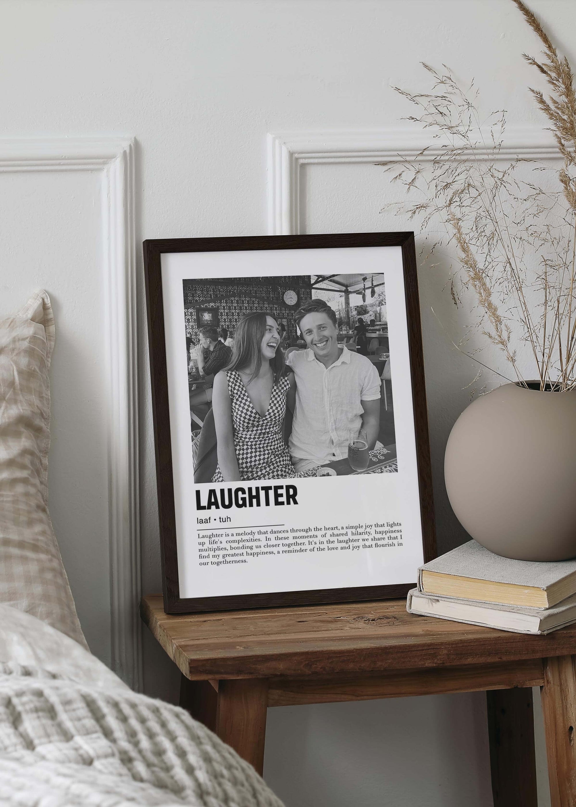 Personalized black and white photo in a black frame of a couple laughing. The custom artwork has a caption that reads Laughter. The black frame is on a small bedside table 