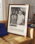 Personalized black and white photo in a walnut frame of a couple laughing. The custom artwork has a caption that reads Laughter. The walnut frame is on a sofa.