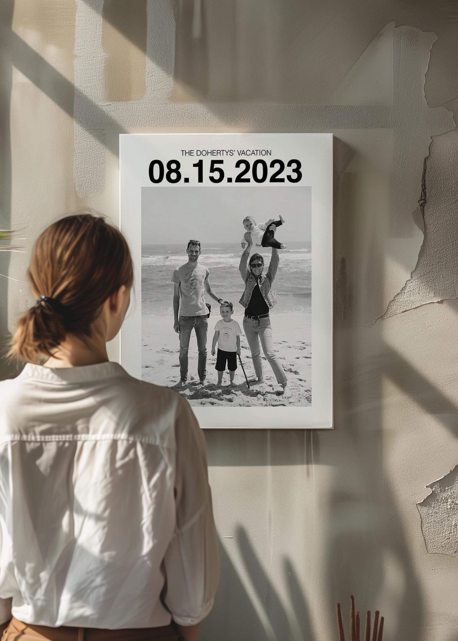 Woman looking at a custom personalized black and white photo of a family on holiday. The canvas has a large custom date at the top and the canvas is mounted on a raw concrete wall.